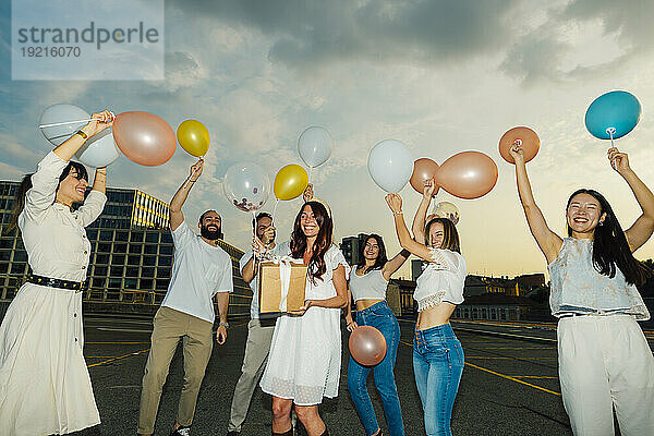 Fröhliche Freunde feiern den Geburtstag der Frau mit Luftballons und Geschenken unter bewölktem Himmel bei Sonnenuntergang