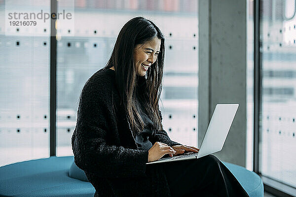 Lächelnde Geschäftsfrau benutzt Laptop auf dem Sitz im Büro