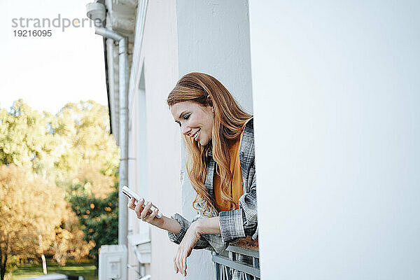Glückliche rothaarige Frau benutzt Smartphone auf Balkon