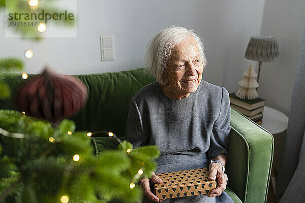 Lächelnde ältere Frau sitzt mit Geschenkbox auf dem Sofa neben dem Weihnachtsbaum zu Hause