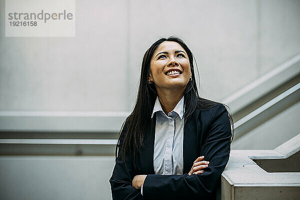 Smiling businesswoman with arms crossed leaning on staircase railing looking up at office