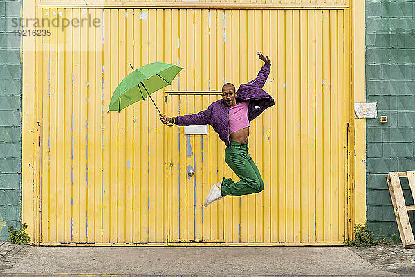 Nicht-binäre Person springt mit Regenschirm vor gelber Fensterladentür