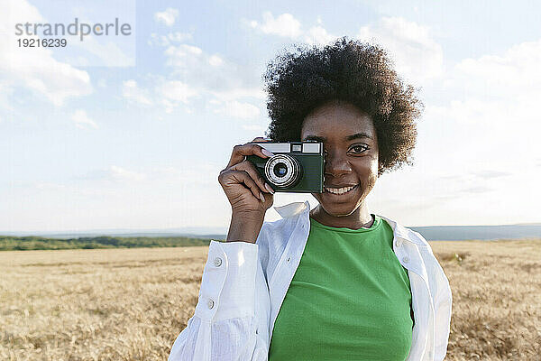 Happy woman with curly hair photographing through camera in field