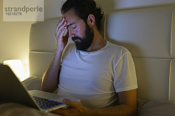 Mann mit Laptop hat zu Hause im Bett Kopfschmerzen