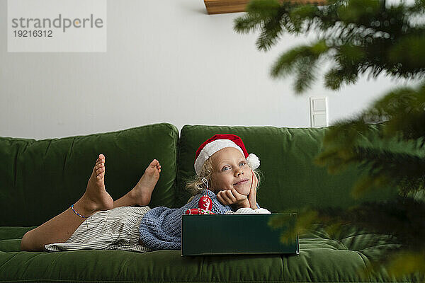 Lächelndes Mädchen liegt auf dem Sofa mit einer Schachtel Weihnachtsschmuck