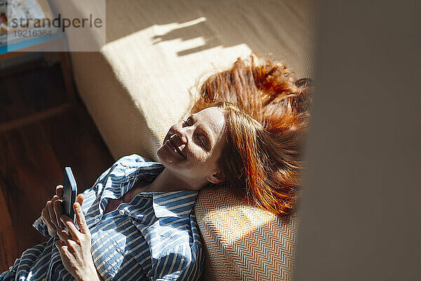 Smiling redhead woman with eyes closed leaning on bed at home