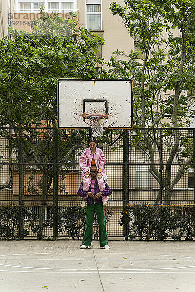 Nicht-binäre Person trägt Frau auf Schultern auf Basketballplatz in der Nähe eines Gebäudes
