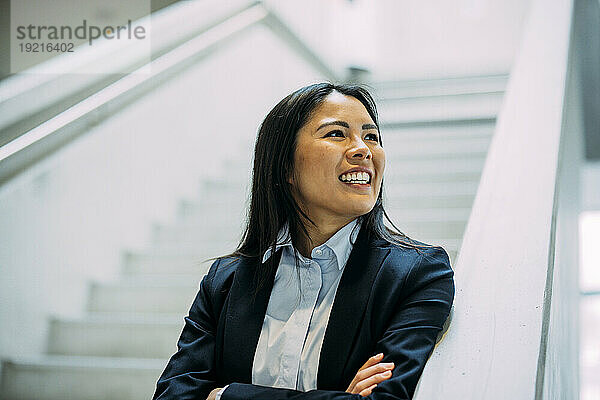 Happy beautiful businesswoman with arms crossed leaning on railing at office