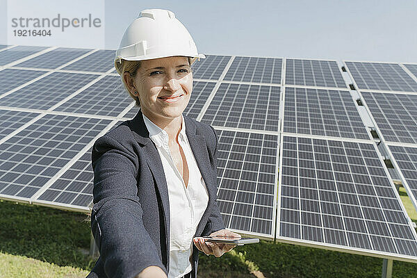 Glücklicher Ingenieur steht mit Smartphone vor Solarpaneelen