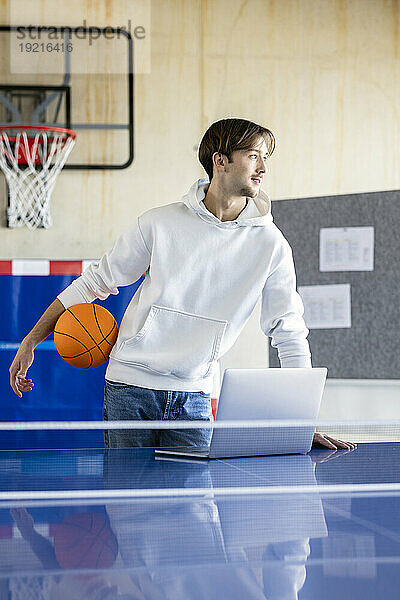 Junger Auszubildender steht mit Basketball und Laptop im Büro