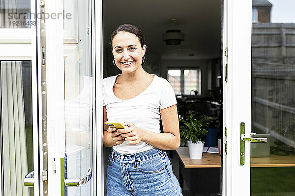 Glückliche Frau steht mit Smartphone im Türrahmen