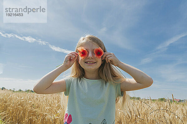 Lächelndes Mädchen hält an einem sonnigen Tag Blumen über die Augen im Weizenfeld