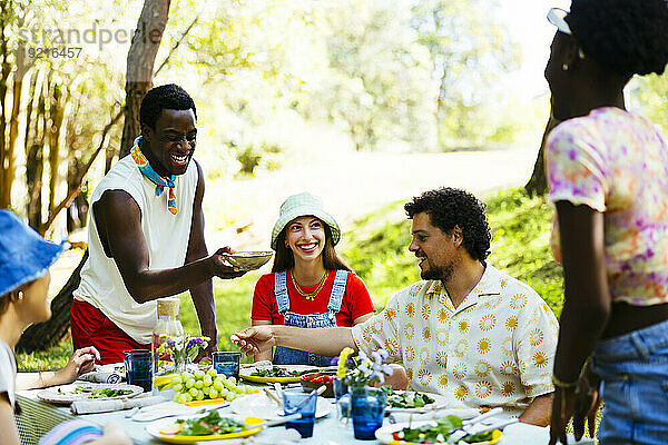 Lächelnder Mann bietet Freunden Essen am Picknicktisch im Garten an