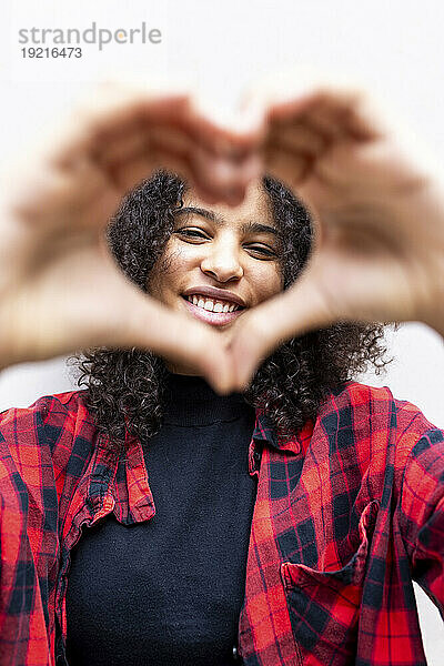 Happy woman with curly hair showing heart sign in front of wall