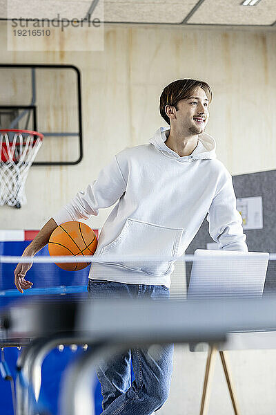 Glücklicher junger Auszubildender steht mit Basketball und Laptop im Büro