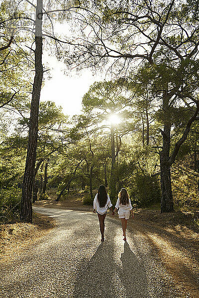 Teenagermädchen hält Händchen mit ihrer Mutter und geht bei Sonnenuntergang auf der Straße im Wald