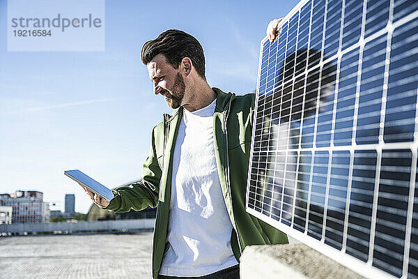 Ingenieur blickt an einem sonnigen Tag auf ein digitales Tablet und hält ein Solarpanel in der Hand