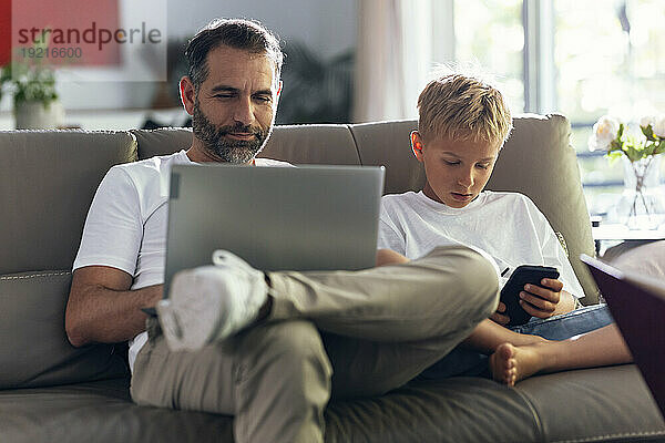 Vater arbeitet am Laptop  Sohn benutzt Smartphone auf Sofa