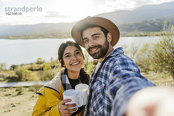 Smiling man taking selfie with girlfriend holding coffee mug