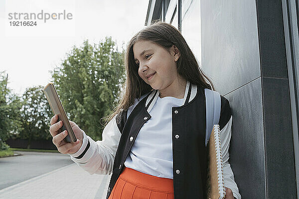 Glückliches Schulmädchen benutzt Smartphone in der Nähe des Schulgebäudes