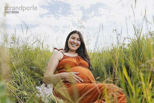 Happy pregnant woman sitting amidst grass under sky