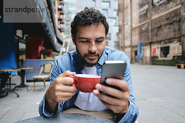 Mann hält Kaffeetasse in der Hand und benutzt Smartphone im Straßencafé