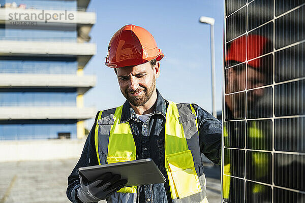 Ingenieur trägt schützende Arbeitskleidung und blickt durch Solarpanel auf ein digitales Tablet