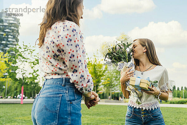 Glückliche Frau mit Freundin hält Geschenkbox und duftenden Blumenstrauß im Park