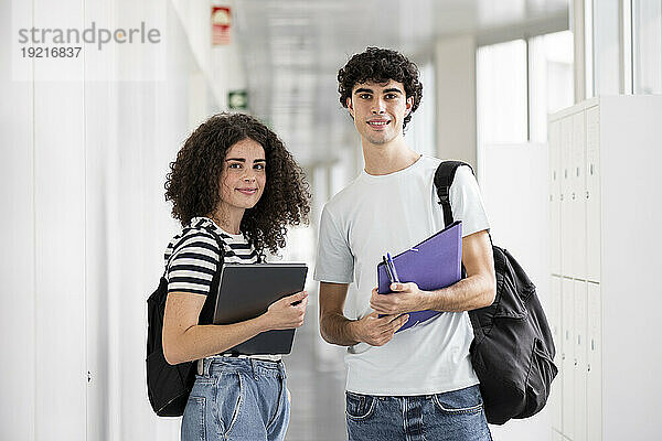 Glückliche Schüler stehen mit Laptop und Aktenordner im Flur
