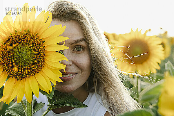 Lächelnde Frau bedeckt Auge mit Sonnenblume im Feld