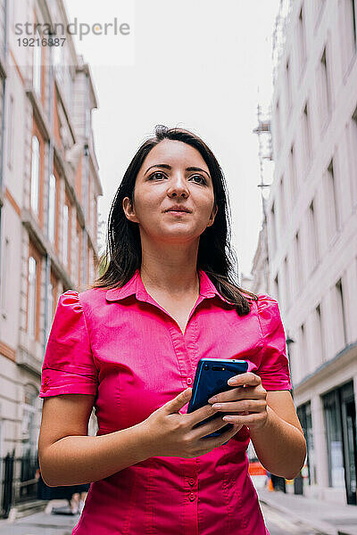 Nachdenkliche Frau hält Smartphone in der Hand und steht inmitten von Gebäuden