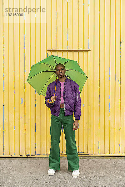 Nicht-binäre Person steht mit grünem Regenschirm vor gelber Fensterladentür