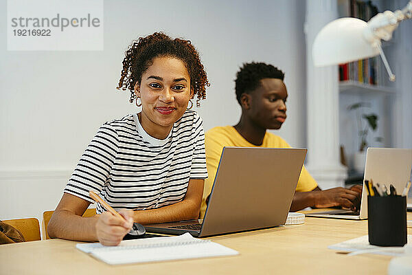 Lächelnder Student mit Freund  der im Hintergrund einen Laptop benutzt