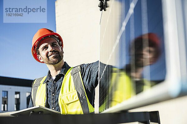 Lächelnder Ingenieur in schützender Arbeitskleidung hält an einem sonnigen Tag ein digitales Tablet in der Hand und untersucht das Solarpanel auf der Terrasse