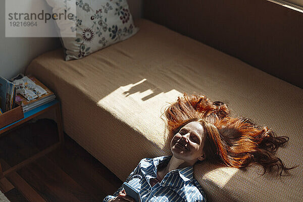 Lächelnde rothaarige Frau lehnt auf dem Bett und hat Sonnenlicht im Gesicht