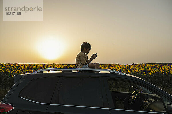 Junge benutzt Smartphone und sitzt bei Sonnenuntergang auf dem Dach eines Autos