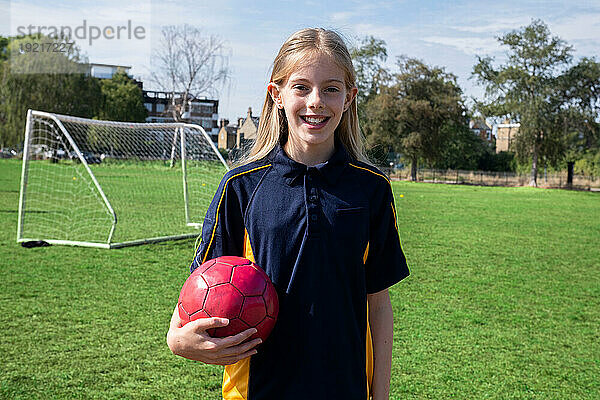 Lächelndes Mädchen mit blonden Haaren hält Sportball auf dem Fußballplatz