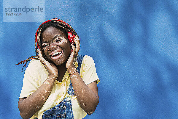 Glückliche junge Frau mit kabellosen Kopfhörern vor blauer Wand
