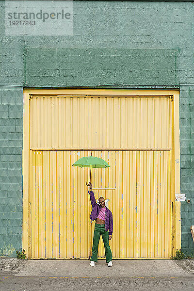 Nicht-binäre Person steht mit Regenschirm vor gelber Fensterladentür