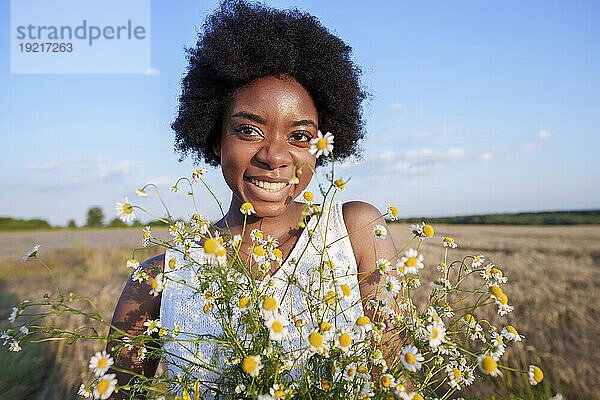 Lächelnde Frau hält an einem sonnigen Tag einen Haufen Gänseblümchen im Feld