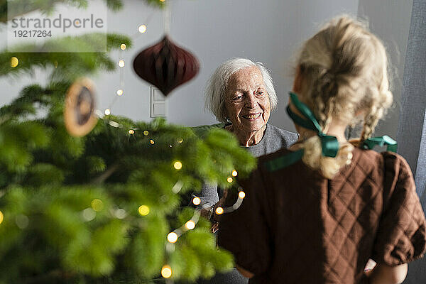 Lächelnde Seniorin mit Enkelin steht zu Hause neben dem Weihnachtsbaum