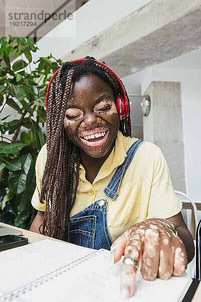 Happy young woman wearing wireless headphones reading book at table
