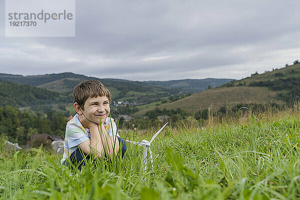 Lächelnder Junge mit Windturbinenmodell auf der Wiese unter bewölktem Himmel