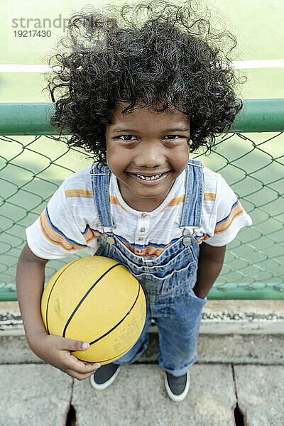 Smiling boy holding basketball near fence at sports court