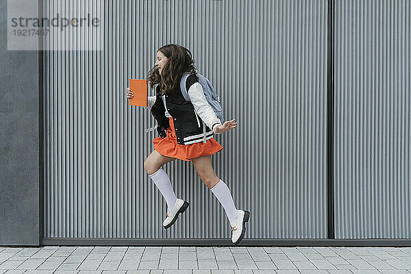 Smiling schoolgirl holding book and jumping in front of wall
