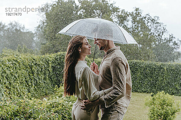 Lächelndes Paar steht mit Regenschirm im Regen im Garten
