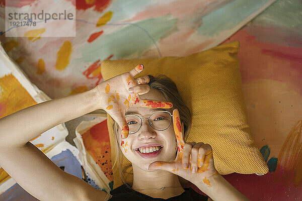 Lächelnde Frau gestikuliert mit Fingerrahmenschild  das auf Gemälden liegt