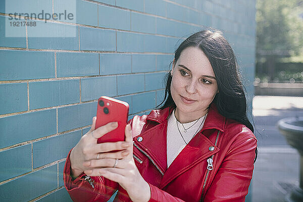 Lächelnde junge Frau macht ein Selfie mit ihrem Smartphone in der Nähe einer Ziegelwand