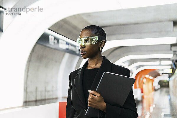 Nachdenkliche junge Frau trägt eine Smart-Brille und steht mit Laptop an der U-Bahn-Station