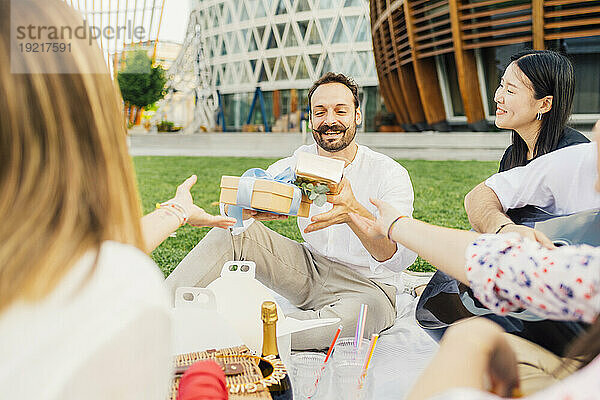 Freunde geben einem glücklichen Mann Geschenke und genießen ein Picknick im Park